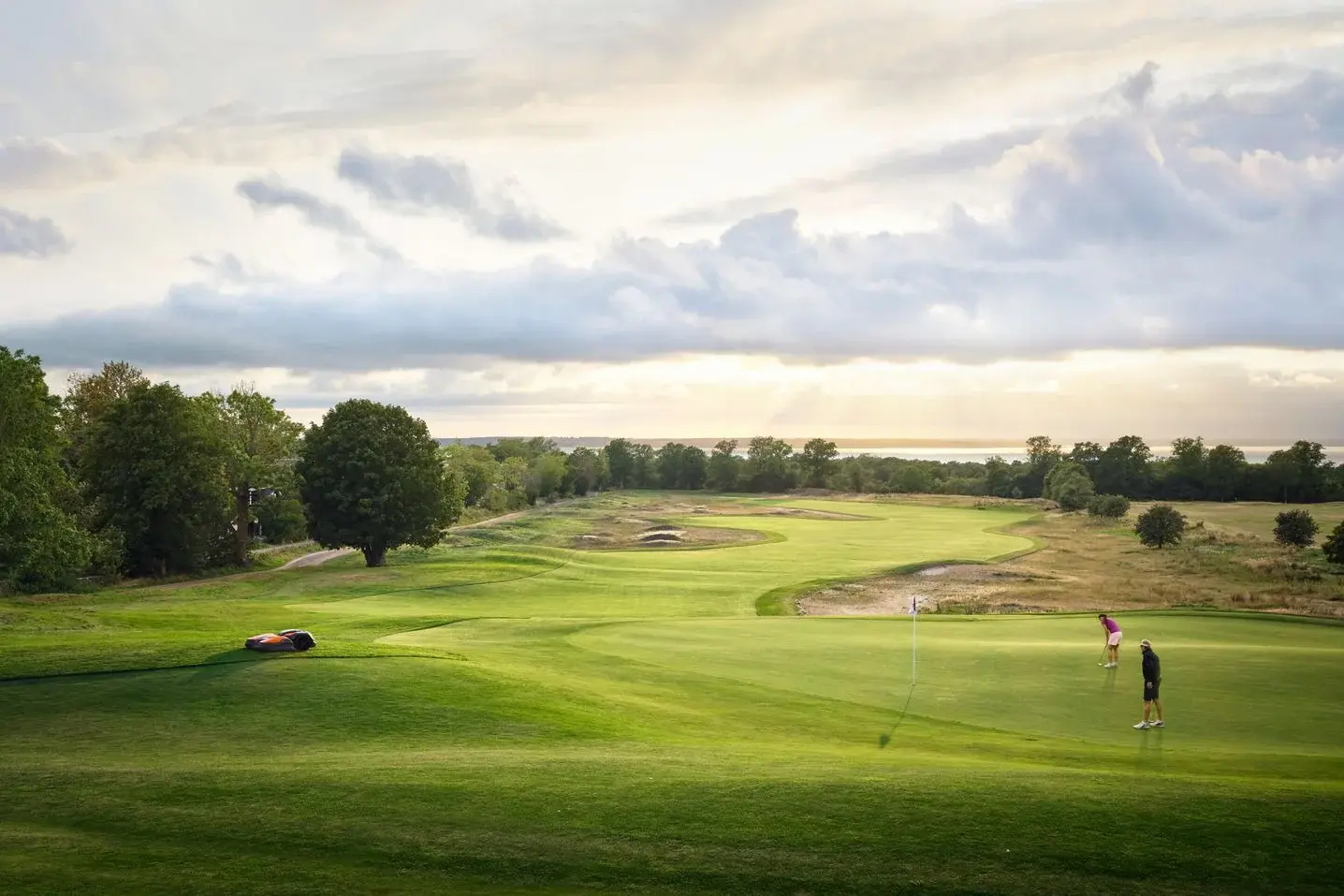 A robotic mower working the fringe of a green at golden hour while two golfers putt, the course rolling out to the sea behind